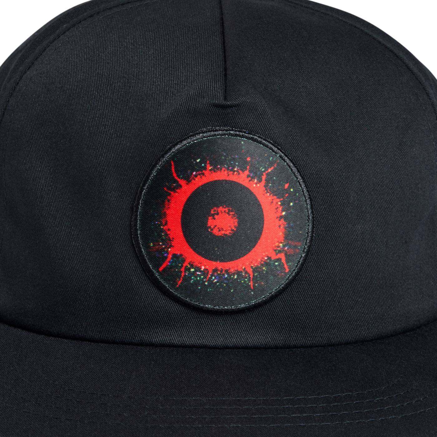 A close-up of a black baseball cap featuring a circular embroidered patch on the front. The patch is black with a bright red splatter design and a small red center, accented with tiny multicolored speckles.