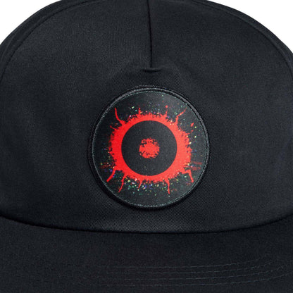 A close-up of a black baseball cap featuring a circular embroidered patch on the front. The patch is black with a bright red splatter design and a small red center, accented with tiny multicolored speckles.
