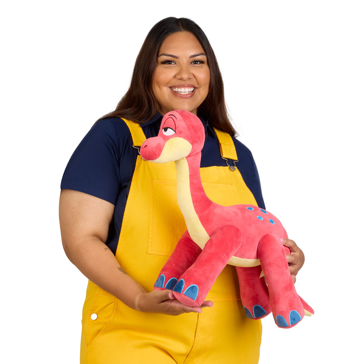 A smiling woman wearing yellow overalls and a navy blue t-shirt holds a long-necked dinosaur plush toy with blue accents.