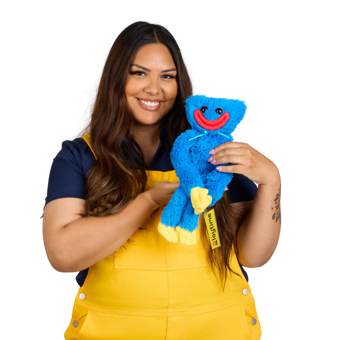 A smiling woman with long dark hair, wearing a navy blue shirt and yellow overalls, holds a blue plush toy with yellow feet and a red lips smiley face.