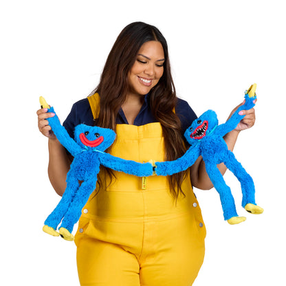 A woman wearing yellow overalls and a navy shirt holds two blue fuzzy monster plush toys, one with a red smile and one with a toothy grin.