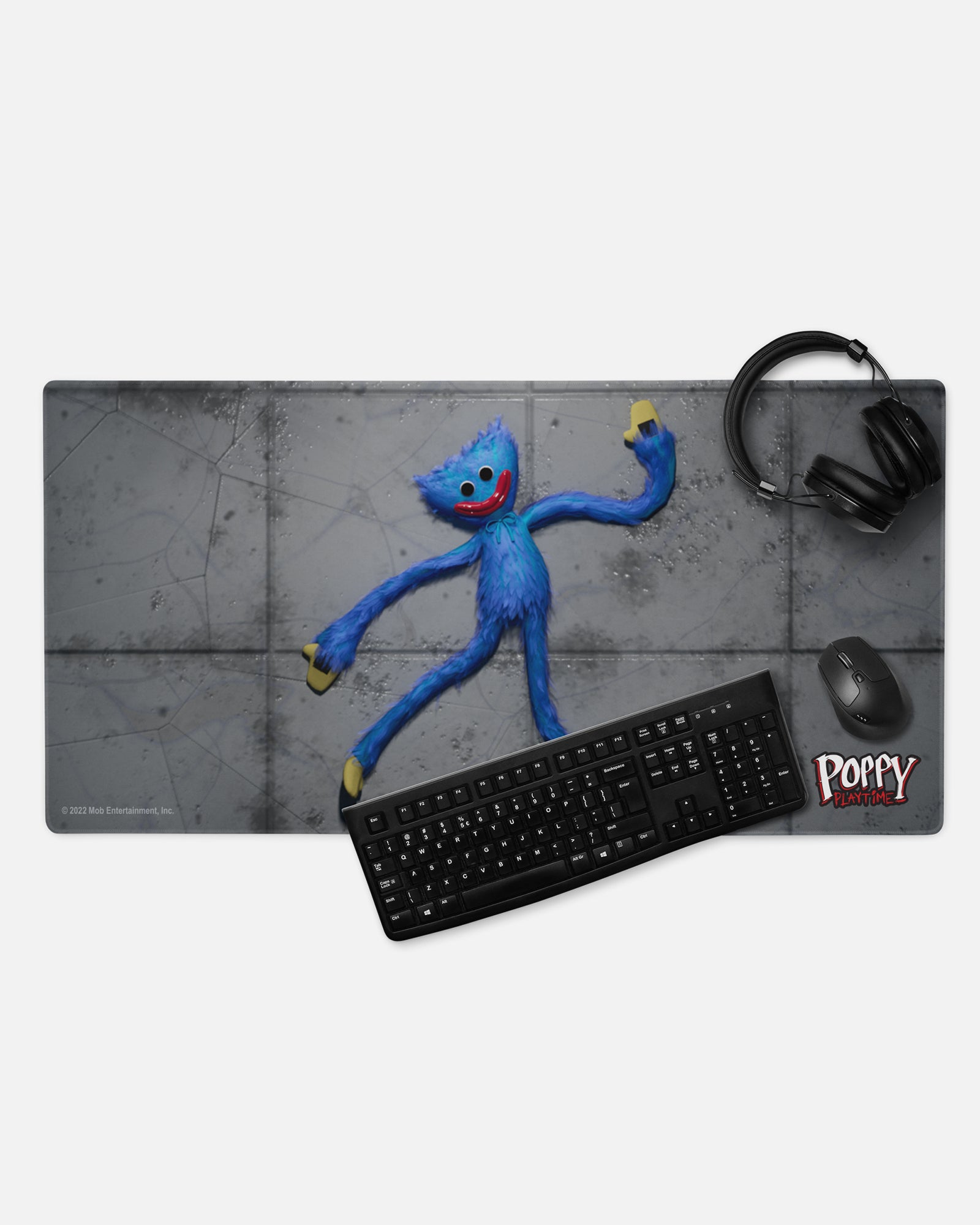 smiling huggy wuggy gamer mousepad with headphones, keyboard, and mouse to show example of gaming set up image: smiling huggy wuggy plush sprawled on concrete floor. text: poppy playtime